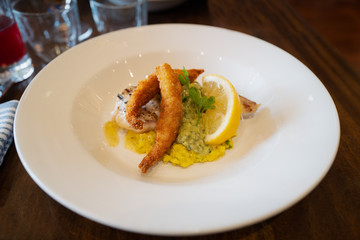 Monkfish with monkfish scampi on a bed of saffron and pesto risotto and a lemon garnish in a white dish on a wooden table.