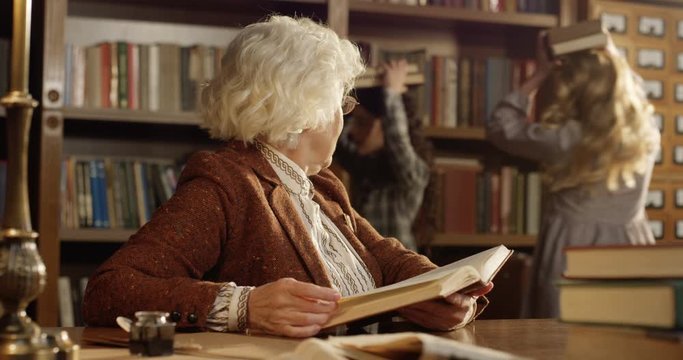 Caucasian old woman in glasses reading book and berating at girls in library at table. Cute kids girls playing with books on background. Grandmother asking granddaughtes not to make noice.