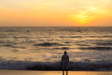 Young man standing at the beach and  looking at amazing sunset by the sea with big foamy waves.