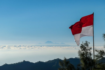 An Indonesian flag waving with the wind seen from the top of Mount Batur. There is Mt Rinjani on Lombok in the back, hiding between clouds. Volcanic landscape of Bali, Indonesia. Island hiking.