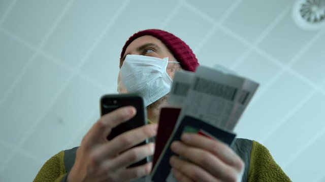 Traveller in protective medical respiratory mask at airport. Man hold passport and boarding pass to cancelled or delayed flight, due to coronavirus outbreak and prevention measures. Scared tourists