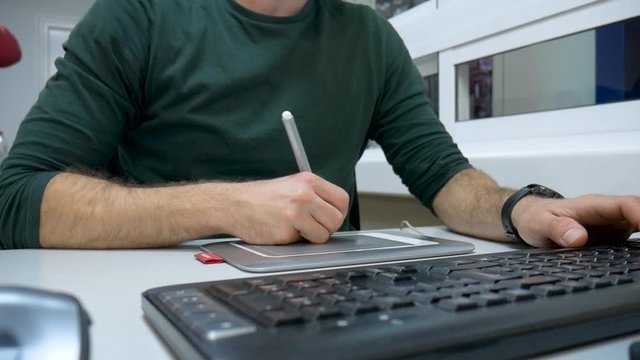 Close up video of a designer drawing with pen on graphic table working in the office. The man wears a smart watch and uses the keyboard. Concept of working at the office.