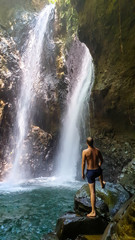 A man in swimsuit standing on the slippery rocks in front of Twin Waterfall, Bali, Indonesia. The waterfall is surrounded by rock from each side. The man enjoyins the beauty of the nature. Adventure