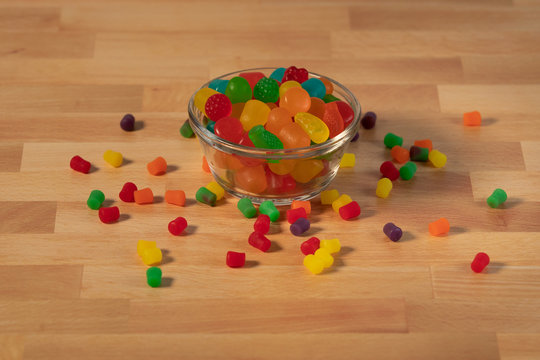 Small glass bowl of multple primary colored candy pieces on butcher wood surface small candy scattered about 