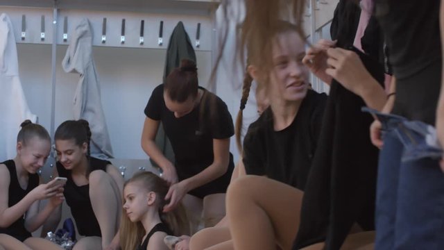 Handheld tracking shot of happy teen and preteen girls changing into dancewear and laughing at something funny on mobile phone before cheerleading practice