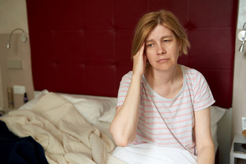 Sad middle-aged woman sitting on the bed in the bedroom. Insomnia, headache, health problems.