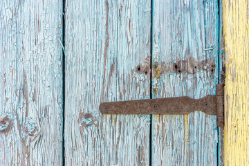Old rusty iron hinge with nails on wooden door with cracked peeling blue and yellow paint close up. Rural background