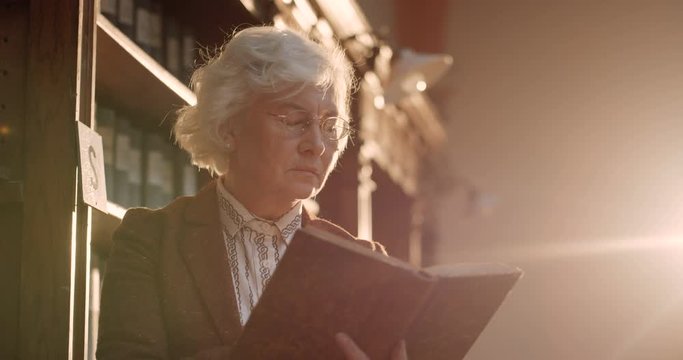 Senior Caucasian gray-haired woman in glasses reading book in ild antique dark library in sun ray. Beautiful female librarian flipping pages in pholiant while working in bibliotheca.