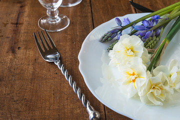 Festive table setting with forged fork and knife and bouquet of daffodils and hyacinths flowers on white plate. Vintage background, close up
