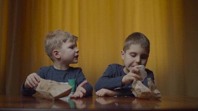 Two siblings in the same clothes eating rice chips at the table at home. Food in paper bag. Boys enjoying eating snacks. 