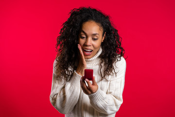 Naklejka premium Amazed african american young woman holding small jewelry box with proposal diamond ring on red wall background. Girl smiling, she is happy to get present, proposition for marriage.