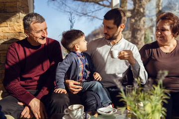 three generations sitting by the table at cafe father grandfather and grandmother and grandson son caucasian family talking small boy and senior man and woman adult in sunny day