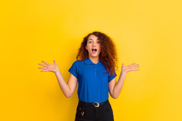 Looks cute, astonished. Caucasian teen's girl portrait on yellow studio background. Beautiful female curly model in blue shirt. Concept of human emotions, facial expression, sales, ad. Copyspace.