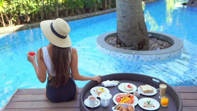 A young woman with a tropical drink in one hand and a buffet of breakfast foods in the other sits poolside with her back to the camera.