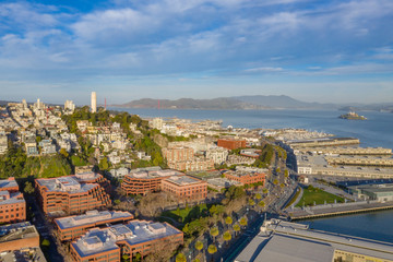 Obraz premium San Francisco, California, daytime, sunrise, aerial view of North Beach with Coit Tower and Golden Gate Bridge visible. Blue sky with clouds and golden light. Embarcadero area in foreground. 
