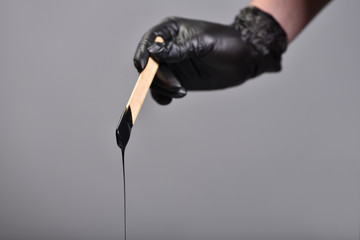 Black wax for depilation on a stick is held by a female hand in a black glove on a gray background