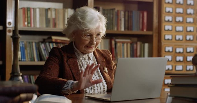 Curious Caucasian old gray-haired woman sitting at table in library and opening lapop computer with surprised eyes. Funny grandmother typing cheerfully like playing on keyboard.