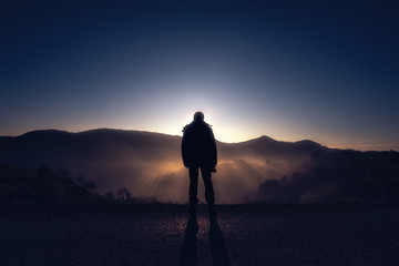 A photographer holds his camera while explores the mountains. A backlit silhouette of a man looking at the sunset.