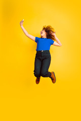 Fototapeta na wymiar Jumping high, taking selfie. Caucasian teen's girl portrait on yellow studio background. Beautiful female curly model. Concept of human emotions, facial expression, sales, ad, education. Copyspace.