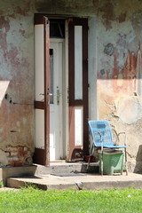 Rusted vintage retro plastic and metal white and light blue chair put on cracked concrete above broken trash can in front of renovated closed and open double doors mounted on dilapidated cracked old s