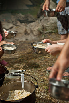 Kids Eating On A Camping Trip. The Food In The Cauldron Is Cooked On Fire. Girl And Boy Holding A Mask. Children Throw Food Together In Plates.