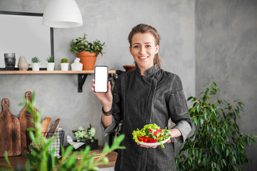 happy young woman shows a smartphone with a white screen on an outstretched hand and holds a plate with chopped vegetable salad in the other hand on the kitchen