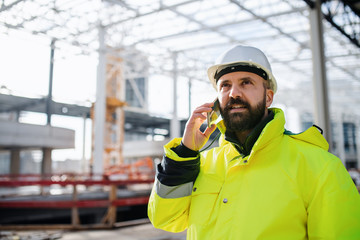 Man engineer standing on construction site, using smartphone.