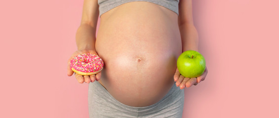 Beautiful young pregnant woman holding green apple and sweet donut in her hand on pink background....