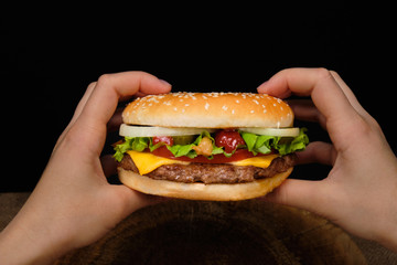 woman holds in her hands from the first person a big burger with meat steak and greens on a black background