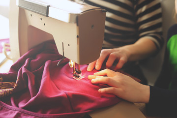 young girl repairs her pink skirt with a small sewing machine under supervision of her mother at home - sustainable fashion concept - obrazy, fototapety, plakaty