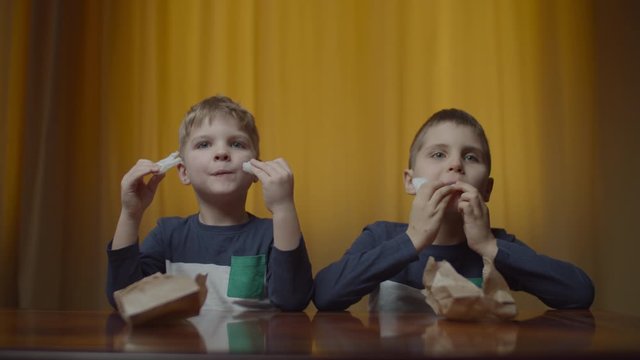 Two siblings in the same clothes eating rice chips at the table at home. Food in paper bag. Boys enjoying eating snacks. 