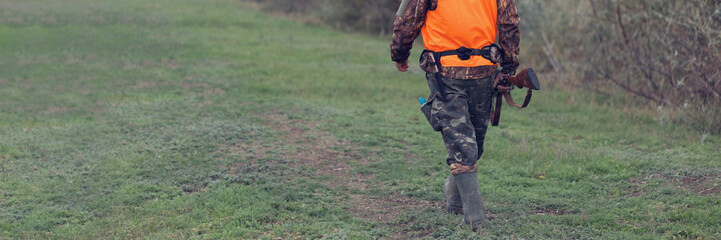 A man with a gun in his hands and an orange vest on a pheasant hunt in a wooded area in cloudy weather. Hunter with dogs in search of game.