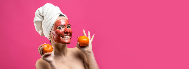 Fototapeta premium Beauty portrait of woman in white towel on head with red nourishing mask on face. Skincare cleansing eco organic cosmetic spa relax concept. A girl stands with her back holding an orange mandarin.