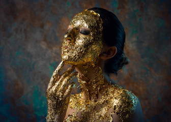 Girl with a mask on her face made of gold leaf. Gloomy studio portrait of a brunette on an abstract background.
