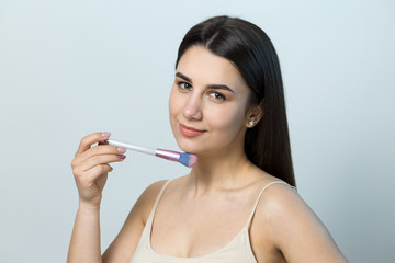 Fototapeta premium Close-up of a young girl in a light top on a white background making a facial make-up. A pretty woman holds a cosmetic brush near her face and smiles.