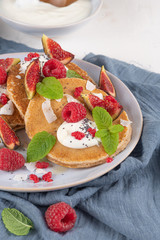 Pancakes with raspberries, figs, yogurt, coconut zest, honey and mint leaves on a plate.