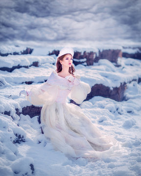 Amazing cute slim young woman in chic fairy tale image in white puffy dress and with crown on her head sits on snow in sunny winter day