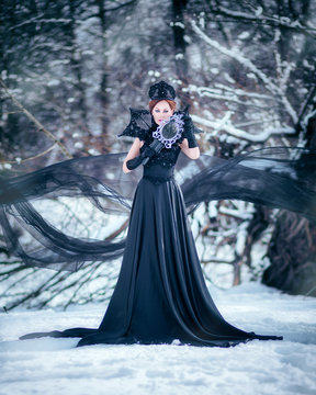 Amazing cute slim young woman in chic fairy tale image in black puffy dress and with crown on her head holds mirror in her hand against background of white snow covered forest