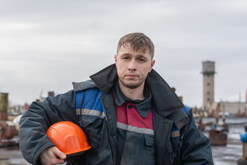 Worker builder on the roof of the factory on the background of electrical equipment on the street. Holds an orange safety helmet under his arm. Dressed in an old, shabby, dirty working uniform.