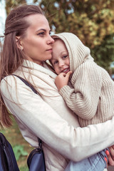 Fototapeta premium Young family, woman mom, holds in her arms child, little son boy, in fall. Walk in city. Emotions of comfort pleasure, smile. Casual warm clothes with hood. Caring for child and parenting.