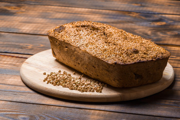 A loaf of freshly baked buckwheat bread lies on a round kitchen board on a brown wooden table. A handful of green buckwheat seeds are scattered nearby. Harmless baking for vegans and vegetarians