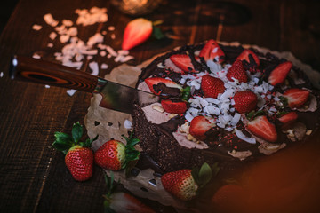 A kitchen knife cutting trough vegan chocolate cake with strawberries, cocnut flakes and raspberry seeds, served on a paper serviette in a soft light of a restaurant, haute cuisine, view from the side