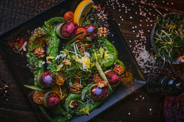 Rich with nutrients appetizer of grilled and oven baked cucumber rolls served with vegan spread, thin sliced vegetables, cherry tomatoes and oil sauce, on a black porcelain dish next to a tiny bowl of