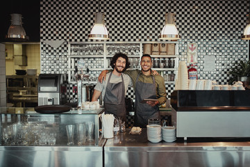 Smiling colleagues standing behind counter in cafe holding digital tablet with hands on each other...