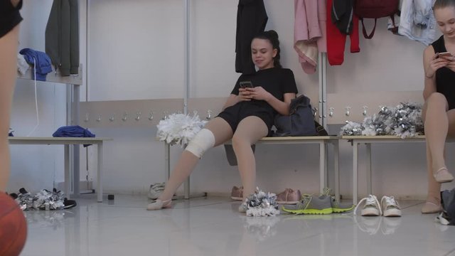 Medium shot of schoolgirl in biketard and gymnastic shoes sitting on bench in locker room and chewing gum while laughing at funny pictures on her phone. Unrecognizable girl with pom-poms practicing