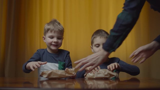 Two siblings in the same clothes eating rice chips at the table at home. Food in paper bag. Boys enjoying eating snacks. 