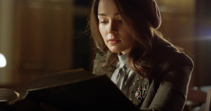 Close up of stylish Caucasian young student girl sitting at table in evening and reading textbook in library. Beautiful woman in hat sitting with old book at desk while studying in bibliotheca.