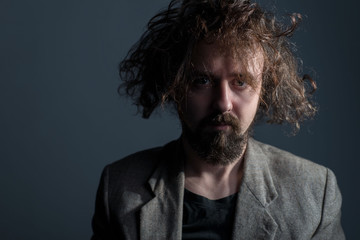Obraz premium Studio dramatic portrait of a young bearded guy, thirty years old, with curly dirty greasy hair and a jacket. On a gray background. The concept of not washed hair. Dirty hair.