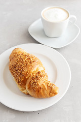 Fresh french croissant on a white plate and a white cup of coffee with foam nearby. Classic french breakfast. Brunch Dessert. Vertical orientation