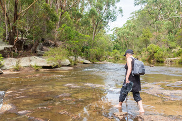 Senior woman with backpack wading through stream holding hiking shoes (selective focus)